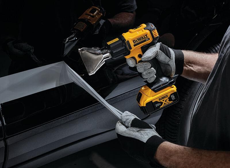 A person wearing gloves uses a DEWALT DCE530B cordless heat gun to apply or remove a film or wrap from the side of a vehicle.