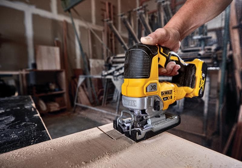 A hand holding a DEWALT DCS334B cordless jigsaw cutting through a piece of wood in a workshop.