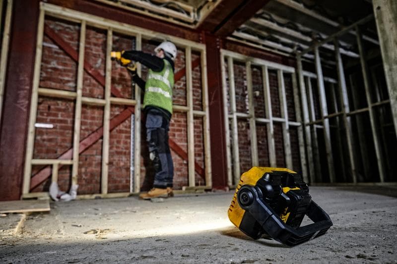 A DEWALT DCL077 work light is placed on the floor of a construction site, illuminating the area while a person wearing