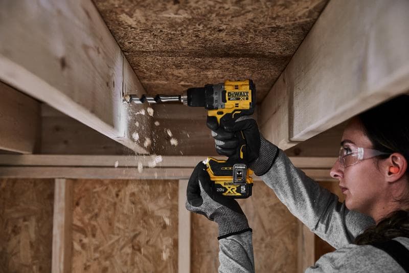 A person wearing gloves is using a DEWALT DCD800P1 20V cordless drill to bore a hole into a wooden beam indoors, with wood shavings falling from the drilled area. The person's face is blurred for privacy.
