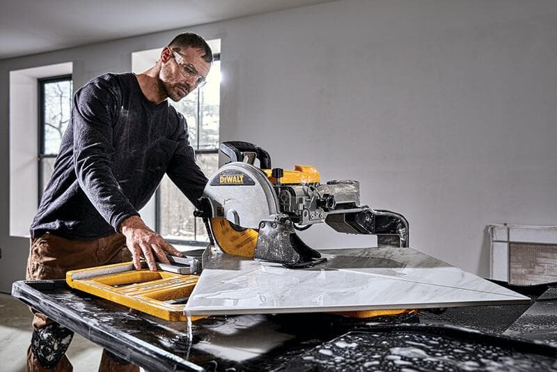 A person using a DEWALT tile saw (model D36000S) to cut a large ceramic tile in a well-lit indoor workspace. The person's face is blurred for privacy.