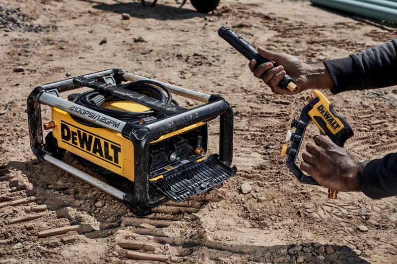 A DEWALT portable pressure washer with visible hoses and attachments is being assembled outdoors on a dirt surface. A person is holding a spray nozzle and connecting it to the machine.
