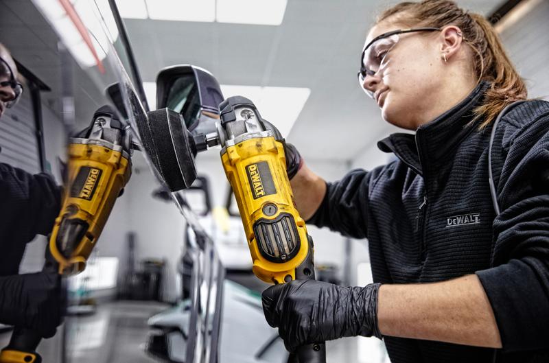 A person wearing black gloves and a black jacket is using a yellow DEWALT DWP849X polisher on the surface of a vehicle, with their face blurred.