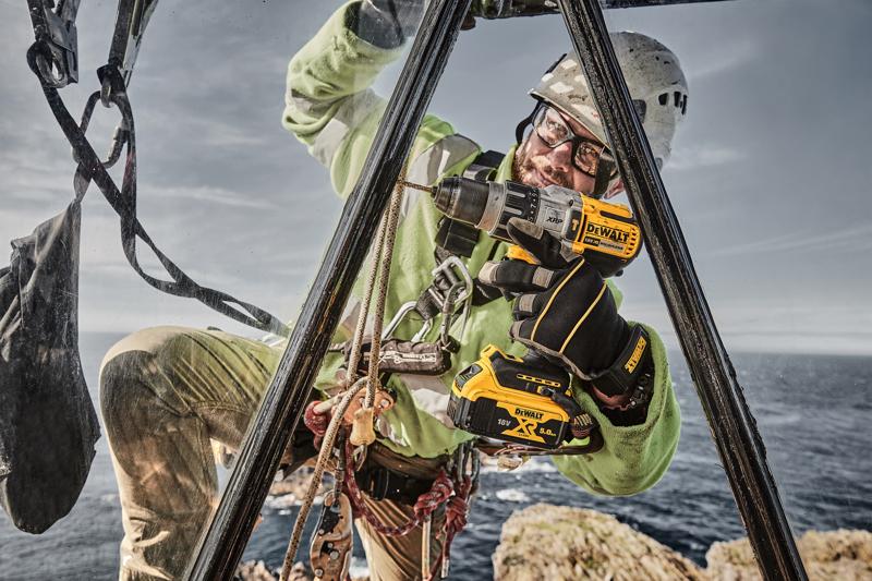 A worker equipped with safety gear uses a DEWALT DCD996P2 cordless power drill outdoors, positioned on a metal structure near a cliff with the sea in the background.