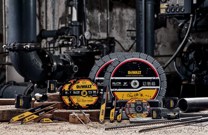 A variety of DEWALT branded industrial tools and accessories, including circular saw blades, drill bits, reciprocating saw blades, and cutting wheels, are displayed on a workbench in a workshop setting with industrial pipes and machinery in the background.