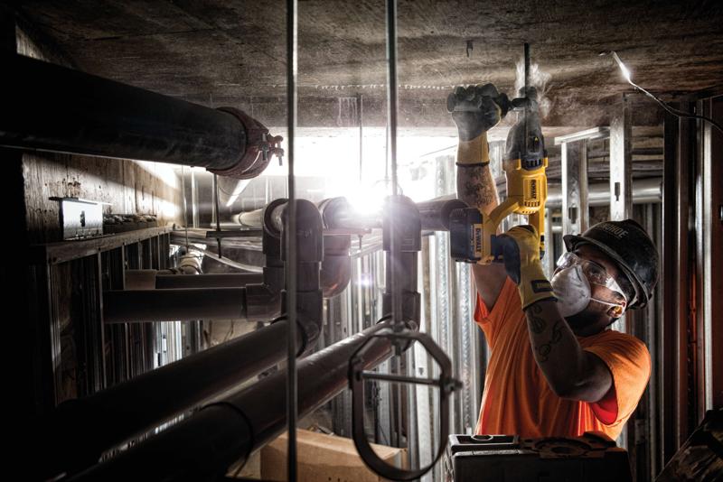 A person wearing a hard hat and safety gloves is using a yellow DEWALT rotary hammer drill to work on a concrete ceiling inside an industrial construction site. Pipes and metal structures are visible around the worker.