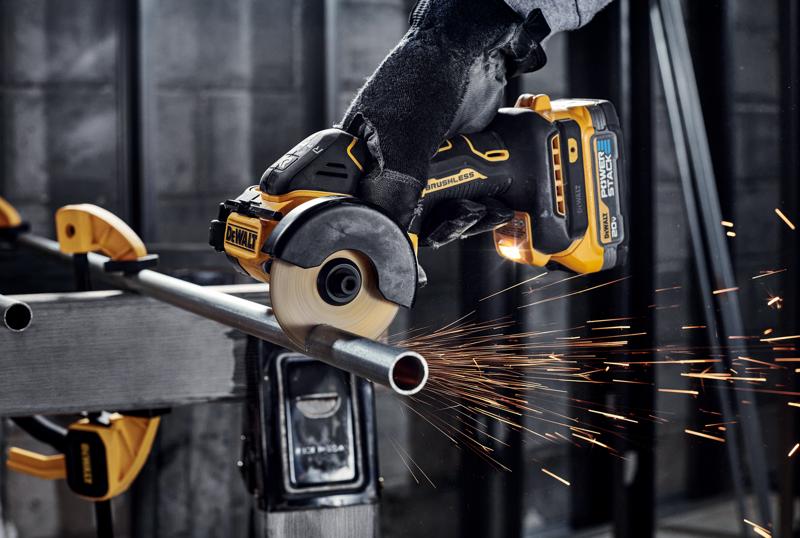 A person wearing gloves uses a DEWALT cordless brushless angle grinder to cut a metal pipe, producing sparks in an industrial workshop setting.