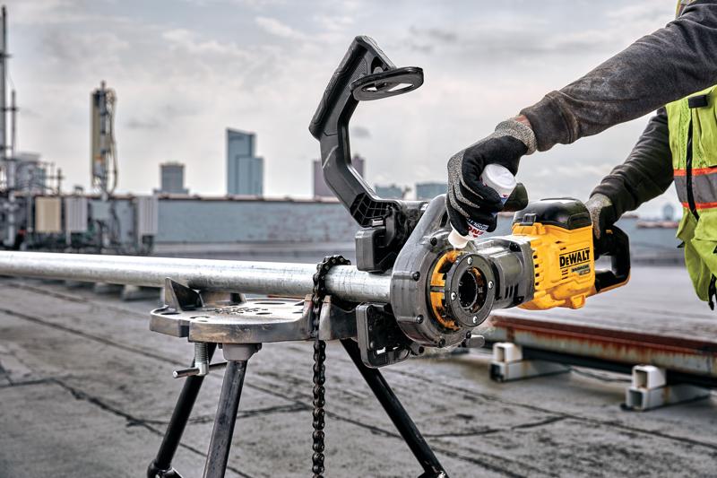 A person using a DEWALT pipe threading machine to thread a metal pipe on a worksite, with a city skyline in the background. The pipe is held in place with a tripod stand and chain, and the worker is wearing gloves and a safety vest.