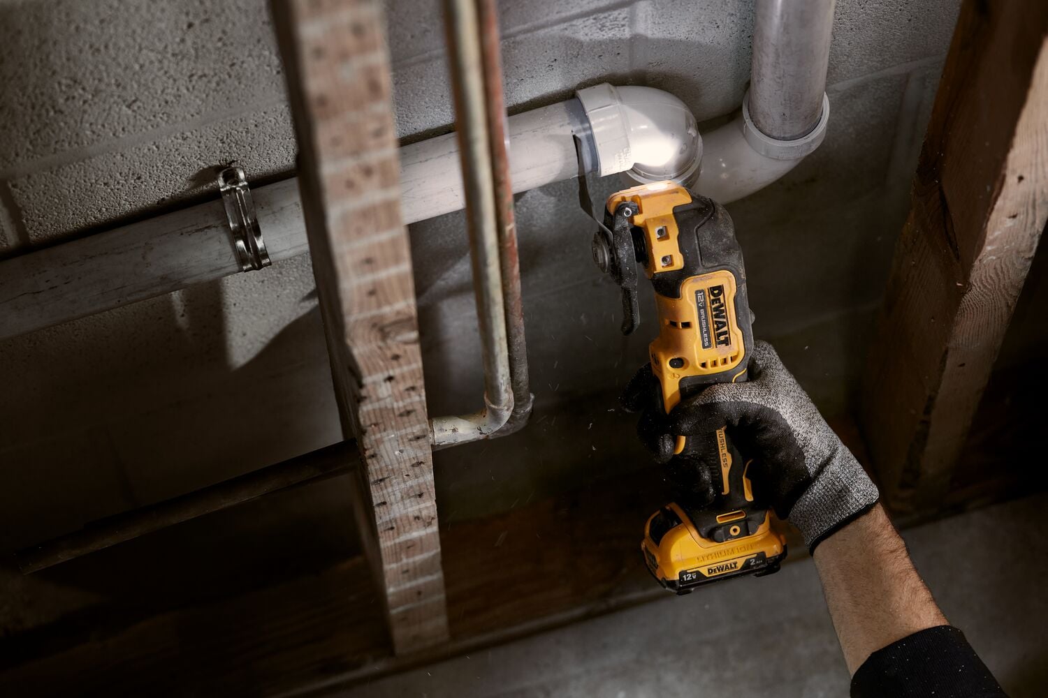 A person wearing a work glove uses a yellow DEWALT cordless oscillating multi-tool to cut a white PVC pipe near a wall in a construction or repair setting.