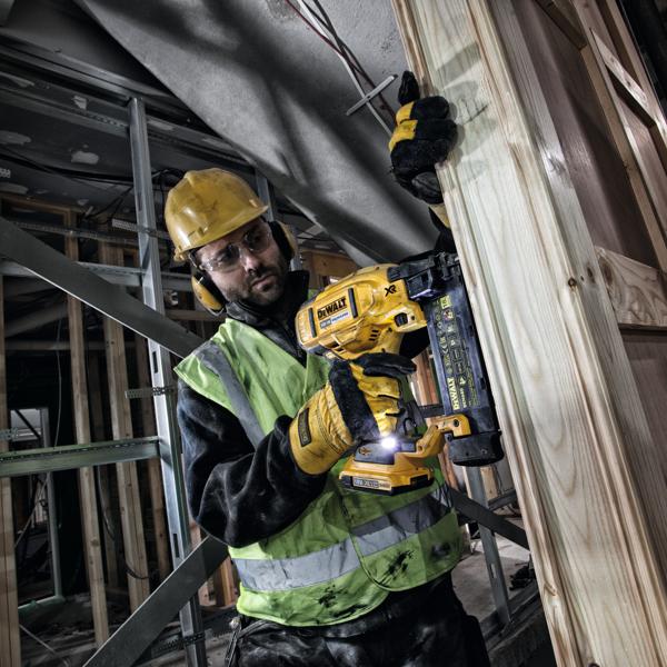 A construction worker wearing a yellow hard hat, reflective vest, and gloves is using a DEWALT DCN680 cordless nailer to fasten wooden panels at a building construction site. The nailer is held firmly against the wood, and the worker is surrounded by unfinished structural elements.