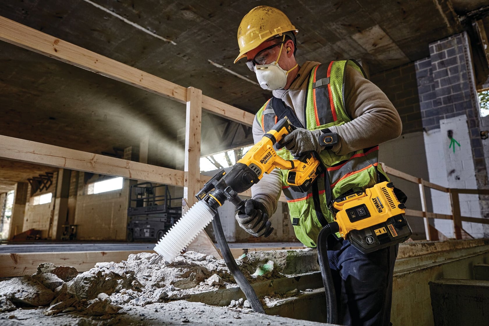 A construction worker wearing a yellow safety helmet and reflective vest operates a DEWALT cordless rotary hammer drill with a dust extraction attachment at a building site. The worker is drilling into concrete debris.
