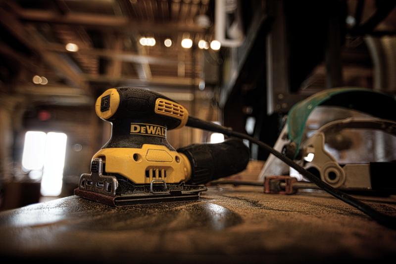 A DEWALT DWE6411K electric sander on a dusty workbench in a workshop, with a power cord visible and various tools in the background.