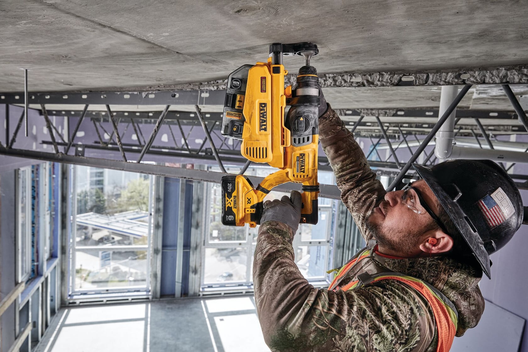 A worker in safety gear is using a yellow DEWALT rotary hammer drill with a dust extraction attachment to drill into a concrete ceiling at a construction site.
