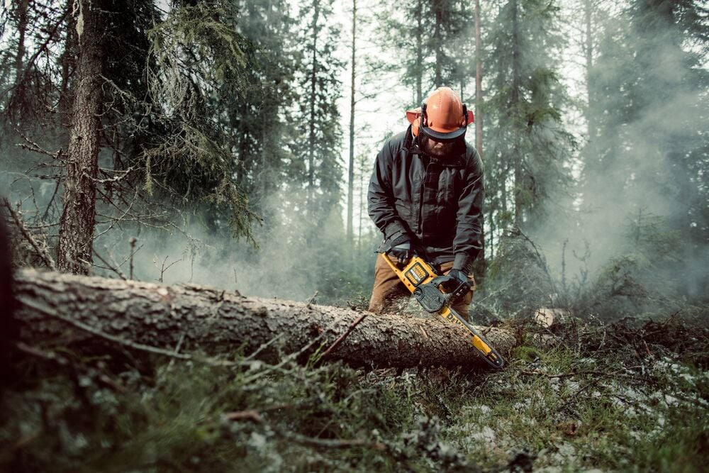 A person wearing protective gear is using a yellow DEWALT chainsaw to cut a fallen tree in a misty forest.