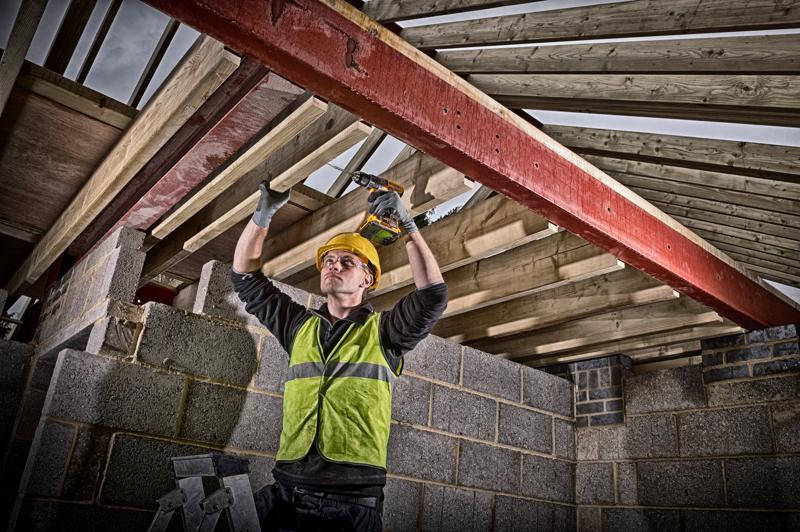 A construction worker wearing a yellow safety helmet and a green reflective vest is using a DEWALT cordless drill to fasten wooden beams to a red steel support inside a building under construction. The walls are made of concrete blocks and wooden rafters are visible above.