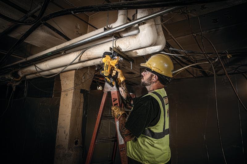 A worker wearing a safety vest and helmet uses a DEWALT portable power tool to cut a metal pipe in an industrial setting. The worker is standing on a ladder and appears to be performing maintenance or installation work on overhead pipes.