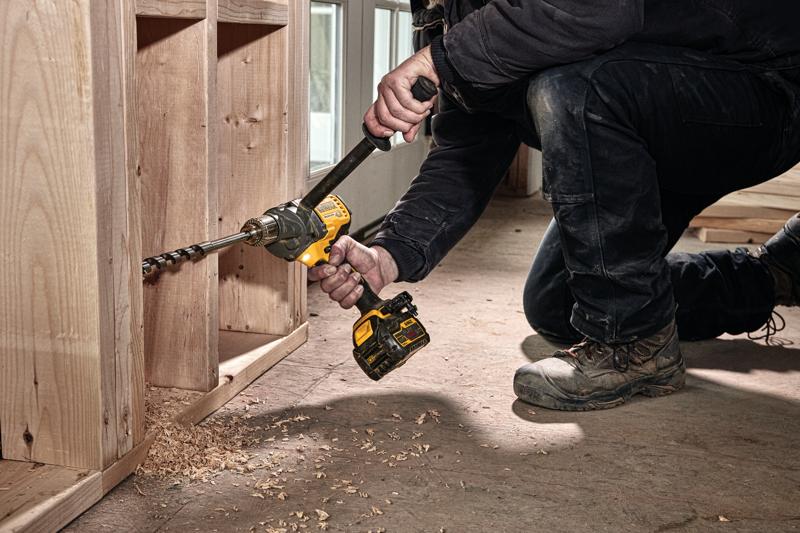 A person kneels on the floor using a DEWALT cordless drill with a long auger bit to drill holes in a wooden cabinet or framing. Wood shavings are scattered on the ground.
