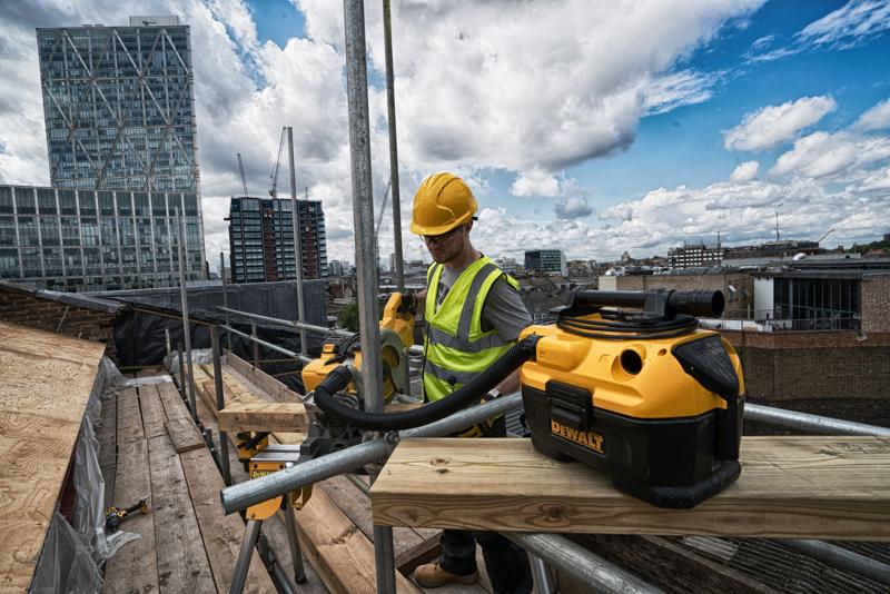 A DEWALT DCV584 vacuum cleaner placed on a construction site, with a worker in a high-visibility vest and helmet operating equipment in the background. Tall buildings and a partly cloudy sky are visible.