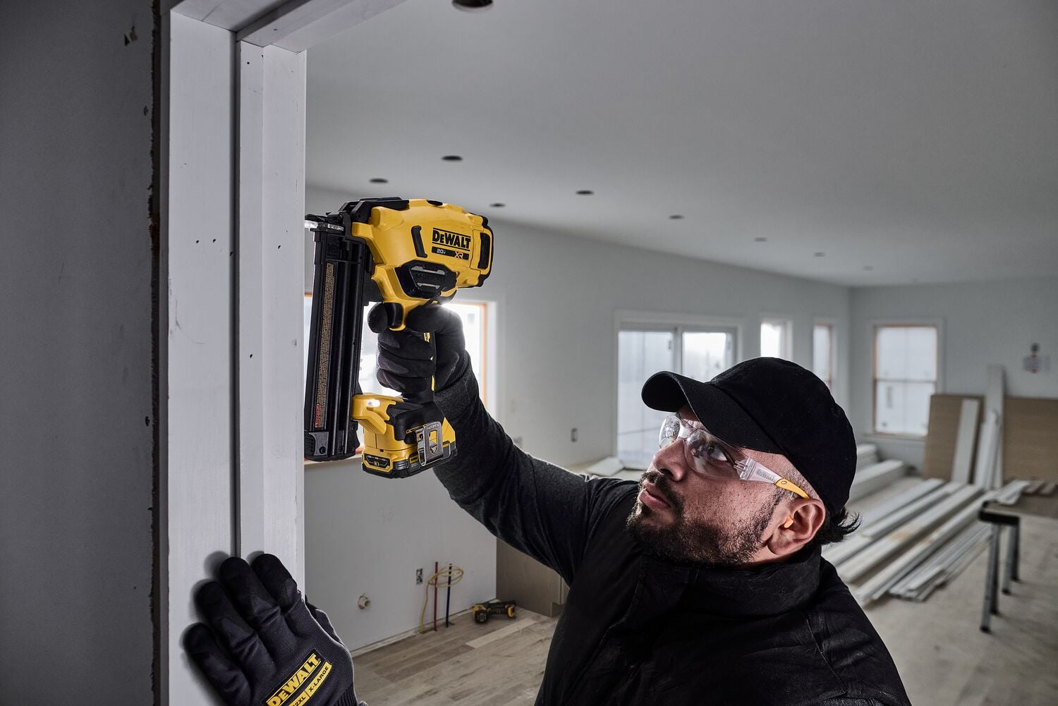 A person wearing gloves is using a DEWALT DCN210 cordless nail gun to fasten trim on a doorway inside a room under construction. The person’s face is blurred and the room has visible building materials and unfinished walls.