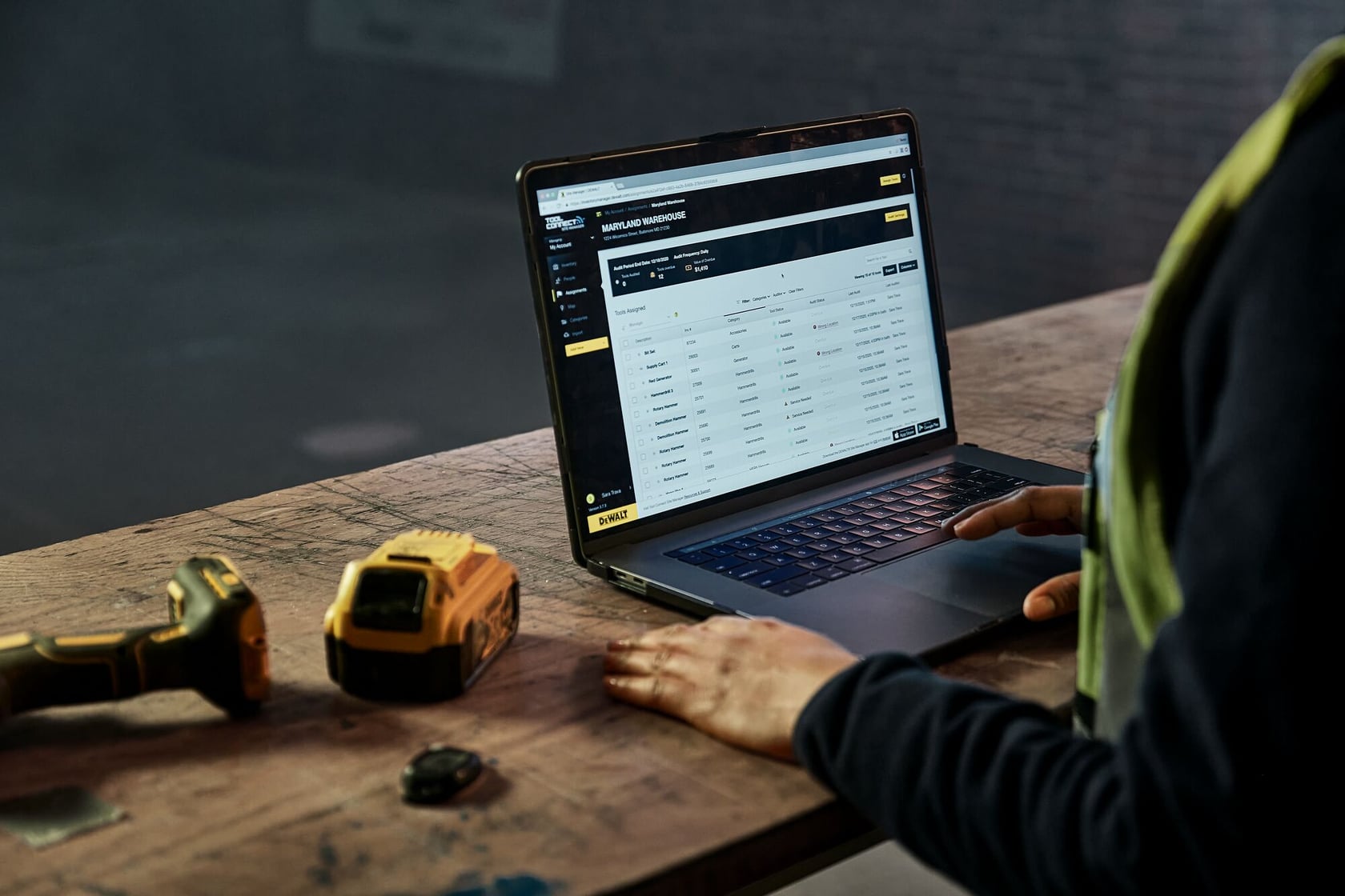 A person using a laptop on a wooden workbench, displaying a DEWALT inventory management interface on the screen. On the bench are a DEWALT cordless tool and a yellow battery pack.