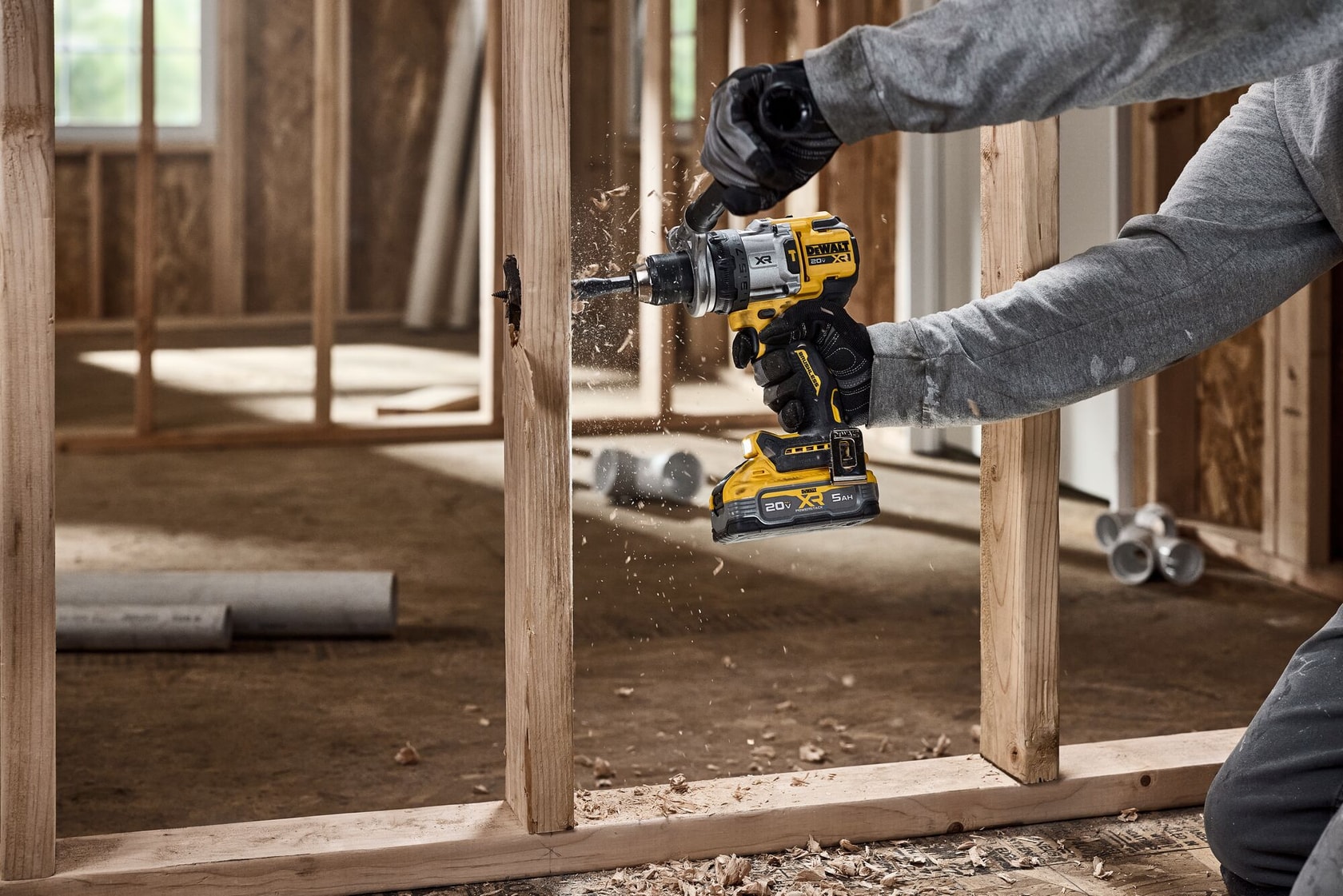 A person wearing work gloves uses a yellow DEWALT cordless drill to bore a hole in a wooden stud inside a construction site.
