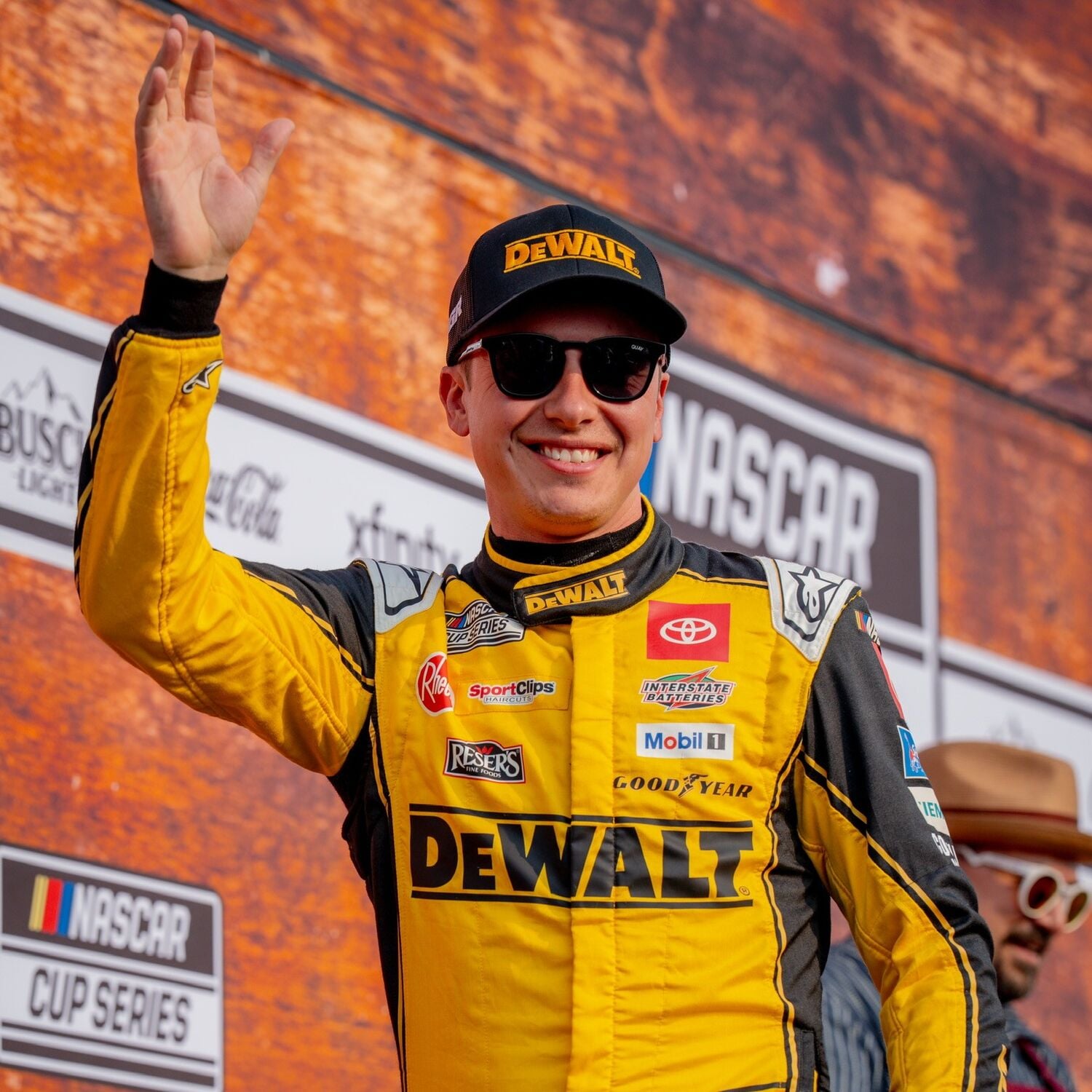A person wearing a yellow DEWALT racing suit and cap, waving on stage at a NASCAR Cup Series event. The background includes NASCAR and Nashville signage.