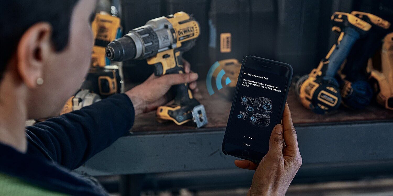 A person uses a smartphone to connect wirelessly with a DEWALT power drill on a workbench, with other DEWALT tools visible in the background.