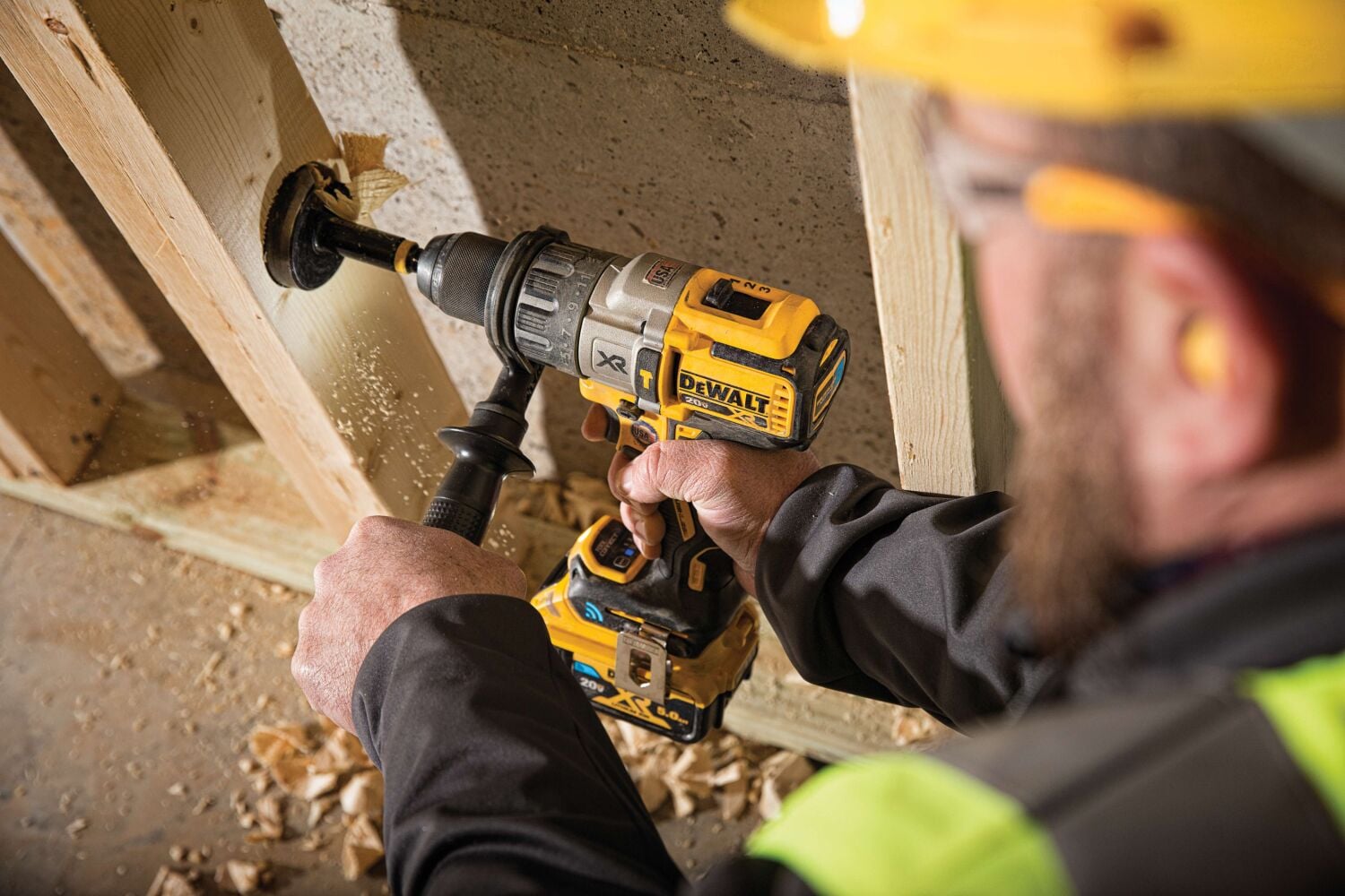 A person wearing safety gear uses a DEWALT cordless drill to bore a hole in a wooden stud on a construction site. Wood shavings are visible around the hole.